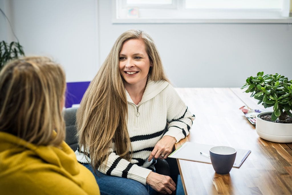A woman with long hair smiles while talking to a friend in a cozy indoor setting, with a plant and notes on the table.