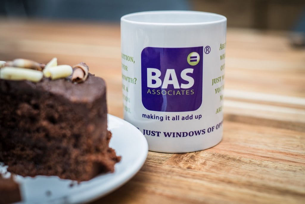 Close-up of a BAS Associates mug next to a slice of chocolate cake on a wooden table.